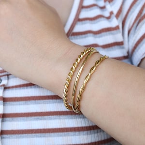 Op de afbeelding: Drie gouden armbandjes, twee met een gedraaid touwontwerp en een met een glad, rond ontwerp.