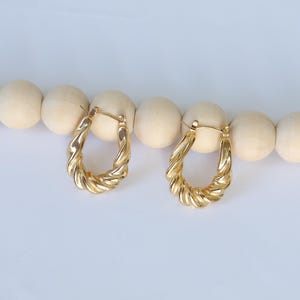 May include: A pair of gold-colored, twisted hoop earrings. The earrings have a textured, rope-like design and a classic oval shape. They are displayed on a string of light-colored wooden beads against a white background.