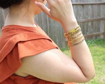 Op de afbeelding: Een persoon draagt een roestkleurig, gerimpeld topje met een gouden kettingarmband en meerdere gouden armbandjes.