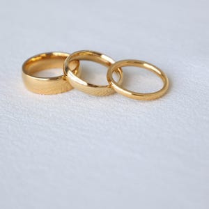 May include: Three gold-colored wedding bands of different sizes, arranged in a row on a white surface.