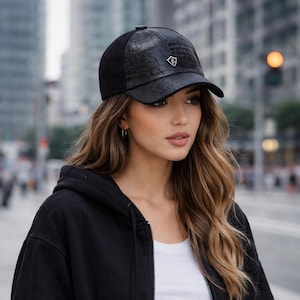May include: Black baseball cap with a textured, faux-crocodile leather front panel and a curved brim. The cap features a small, silver emblem on the front and a black fabric back. The cap is worn by a person in a black hoodie.