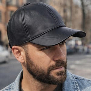 May include: A black leather baseball cap with a curved brim. The cap has a structured design with visible stitching and a button on top. The hat is worn by a person in a denim jacket.