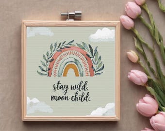 May include: A framed cross-stitch design featuring a rainbow with green leaves and the text "stay wild, moon child."