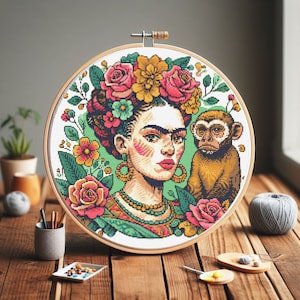 Peut inclure: Un cerceau de broderie coloré avec un portrait de Frida Kahlo portant une couronne de fleurs et un singe sur son épaule. L'arrière-plan est vert avec des fleurs et des feuilles.