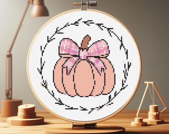 May include: A cross-stitch embroidery hoop with a pink pumpkin with a pink bow in the center. The pumpkin is surrounded by a wreath of leaves.