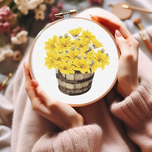 May include: A cross-stitch embroidery hoop with a design of a wooden bucket filled with yellow flowers.