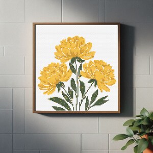 May include: A framed cross-stitch artwork featuring three large yellow flowers with green stems and leaves against a white background. The artwork is in a brown wooden frame and is displayed on a white tiled wall.
