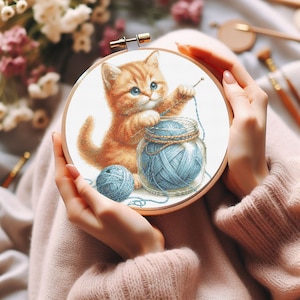 Puede incluir: Un aro de bordado de punto de cruz con fondo blanco que presenta un lindo gatito naranja jugando con una bola de lana azul en un frasco de vidrio. El gatito sostiene una aguja en su pata.