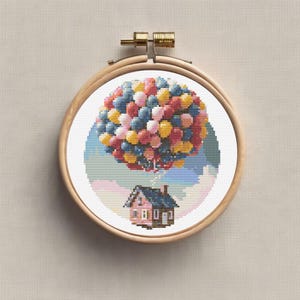Peut inclure: Broderie au point de croix d'une maison soulevée par un bouquet de ballons multicolores. Les ballons sont bleus, roses, jaunes et rouges. La maison rose a un toit bleu, dans un cercle de broderie en bois.