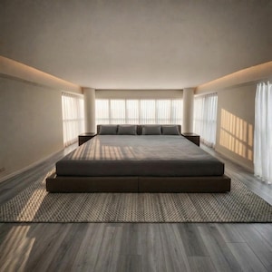 Puede incluir: Una cama de plataforma baja con un colchón gris oscuro y cuatro almohadas rectangulares a juego. La cama está centrada sobre una alfombra gris grande y texturizada en una habitación con paredes claras, suelo de madera y ventanales.