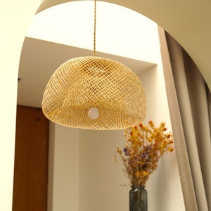 Op de afbeelding: Een geweven rotan hanglamp hangt aan het plafond in een kamer met witte muren en een bruin gordijn. De lamp is rond en heeft een witte gloeilamp erin.
