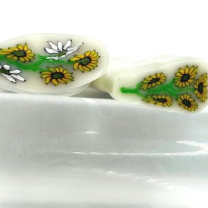 May include: Two white, oblong-shaped beads with a floral design. Each bead features yellow sunflowers and white daisies with green stems. The beads are set against a white background.