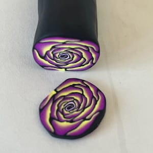 May include: A close-up of a polymer clay cane with a purple and yellow rose design. The cane is cut into a round slice, showing the intricate details of the rose.