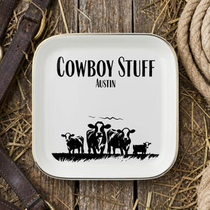 May include: A square white ceramic trinket dish with a gold-toned rim. The center features black text reading COWBOY STUFF and AUSTIN above a black graphic illustration of four cows standing in a field of grass, showcasing a rural aesthetic.