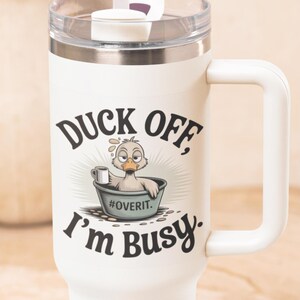 Pode incluir: Caneca térmica branca com alça e tampa transparente. A caneca apresenta um pato de desenho animado em uma banheira com o texto "DUCK OFF, I'm BUSY". O pato está segurando uma caneca e a hashtag #OVERIT também é visível.