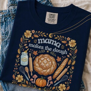 May include: A navy blue t-shirt with a graphic print featuring the text "mama makes the dough" surrounded by illustrations of a braided loaf of bread, a baguette, a jar of flour, rolling pins, whisks, a pumpkin, mushrooms, and autumn leaves.