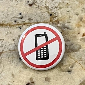 May include: A round white button with a red circle and a black cell phone icon crossed out. The button has a glossy finish and is likely a pin or badge.