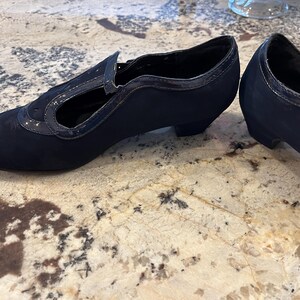 K&ouml;nnte beinhalten: Ein Paar marineblaue Schuhe im Vintage-Stil. Die Schuhe haben eine runde Spitze, einen kleinen Absatz und einen Riemen &uuml;ber dem Fu&szlig;. Der obere Teil besteht aus einem weichen, Wildleder-&auml;hnlichen Material mit gl&auml;nzendem Besatz. Die Abs&auml;tze sind ca. 5 cm hoch.