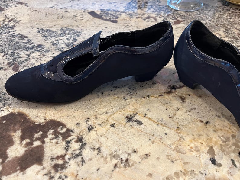 K&ouml;nnte beinhalten: Ein Paar marineblaue Schuhe im Vintage-Stil. Die Schuhe haben eine runde Spitze, einen kleinen Absatz und einen Riemen &uuml;ber dem Fu&szlig;. Der obere Teil besteht aus einem weichen, Wildleder-&auml;hnlichen Material mit gl&auml;nzendem Besatz. Die Abs&auml;tze sind ca. 5 cm hoch.