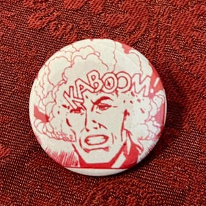 May include: A round, white button with a red comic book style illustration. The image features a surprised face with the word "KABOOM!" in red text. The button is set against a textured, red background.
