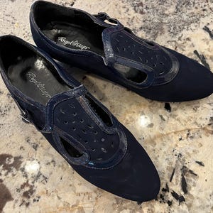 K&ouml;nnte beinhalten: Ein Paar marineblaue Angel Steps Schuhe. Die Schuhe haben ein T-Riemen-Design mit Ausschnittdetails und eine gl&auml;nzende Lacklederbesatz. Sie haben einen Schnallenverschluss und eine abgerundete Spitze.