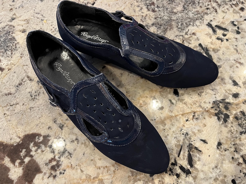 K&ouml;nnte beinhalten: Ein Paar marineblaue Angel Steps Schuhe. Die Schuhe haben ein T-Riemen-Design mit Ausschnittdetails und eine gl&auml;nzende Lacklederbesatz. Sie haben einen Schnallenverschluss und eine abgerundete Spitze.