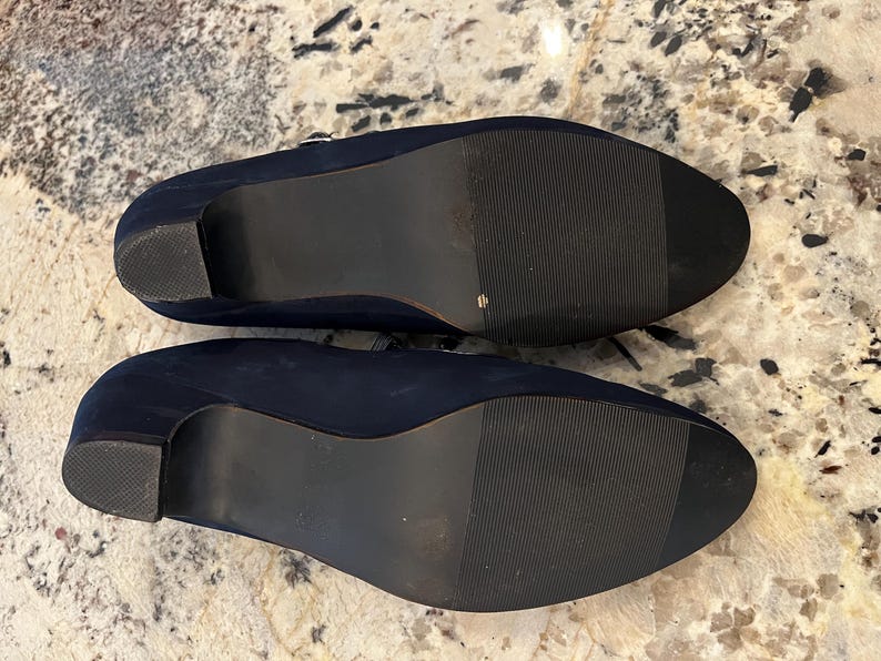 K&ouml;nnte beinhalten: Ein Paar marineblaue Schuhe mit Absatz und dunklen Sohlen. Die Schuhe haben eine abgerundete Spitze und einen kleinen Absatz. Die Sohlen sind mit horizontalen Linien f&uuml;r den Halt strukturiert. Die Schuhe sind wahrscheinlich aus Wildleder oder einem &auml;hnlichen Material.