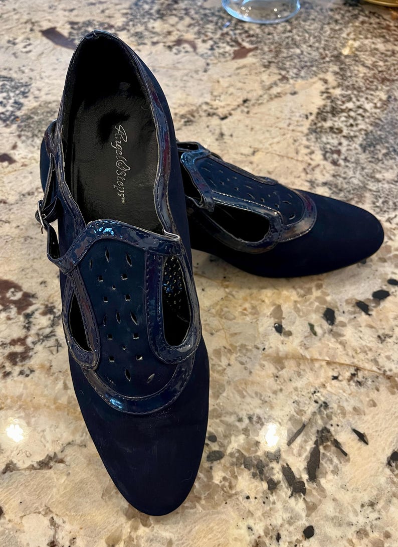 K&ouml;nnte beinhalten: Ein Paar marineblaue Schuhe mit niedrigem Absatz. Die Schuhe haben eine Wildleder-&auml;hnliche Textur an Zehen und Ferse, mit einem gl&auml;nzenden, perforierten Lackleder-Overlay. Die Schuhe haben einen Riemen mit Schnallenverschluss.