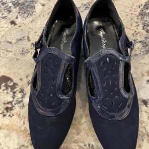 K&ouml;nnte beinhalten: Ein Paar marineblaue Schuhe mit T-Riemen-Design. Die Schuhe haben eine matte Oberfl&auml;che an der Spitze und einen gl&auml;nzenden Lackriemen mit Ausschnitten. Der Markenname "Angel Steps" ist auf der Innensohle sichtbar.