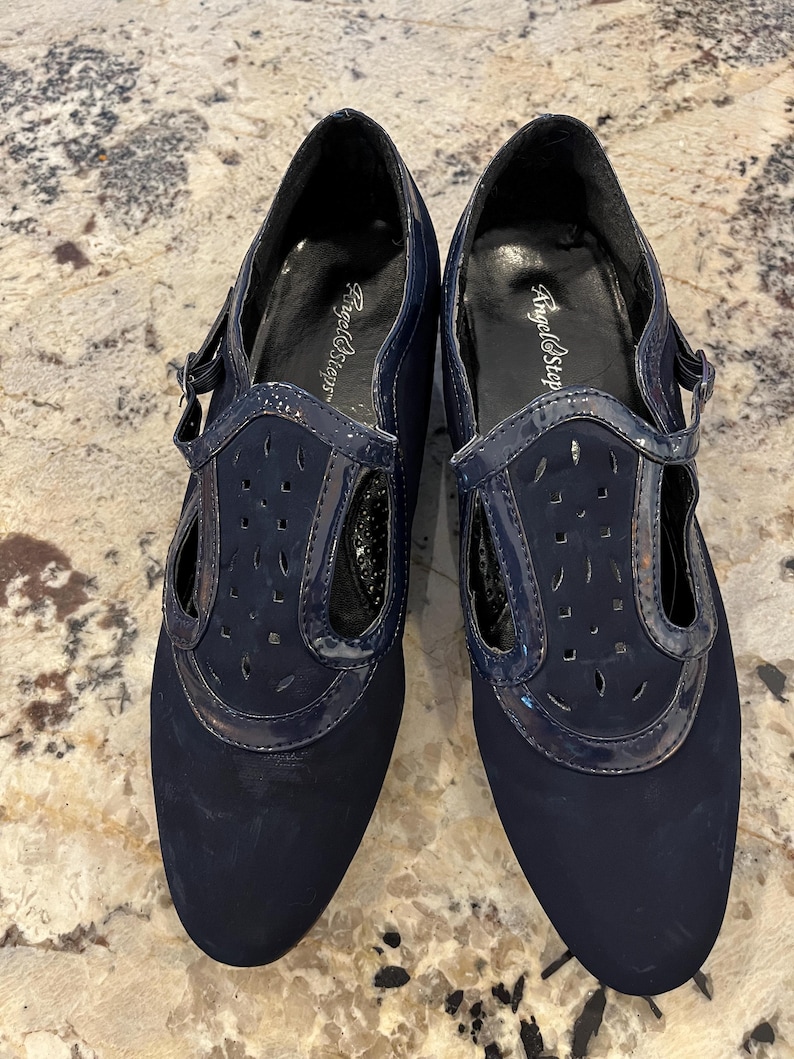 K&ouml;nnte beinhalten: Ein Paar marineblaue Schuhe mit T-Riemen-Design. Die Schuhe haben eine matte Oberfl&auml;che an der Spitze und einen gl&auml;nzenden Lackriemen mit Ausschnitten. Der Markenname "Angel Steps" ist auf der Innensohle sichtbar.