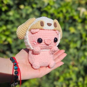 Puede incluir: Un cerdito rosa hecho a mano con ganchillo, que lleva un sombrero beige y blanco con detalles marrón oscuro. El cerdo tiene grandes ojos negros y se sostiene en una mano sobre un fondo verde borroso. Se ve una pulsera roja con cuentas azules y negras.