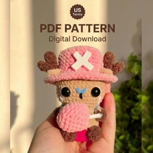Puede incluir: Una figura amigurumi de ganchillo de una criatura color canela con astas marrones, que lleva un sombrero rosa con una cruz blanca. Tiene ojos negros, nariz azul y sostiene una esfera rosa texturizada. Texto: US Terms, PDF PATTERN, Digital Download.