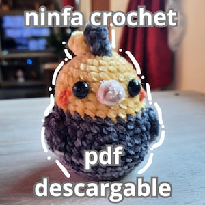 May include: A handmade crochet bird in yellow, gray, and orange. The bird has black eyes and a small white beak. The text "ninfa crochet" is above the bird, and "pdf descargable" is below it.