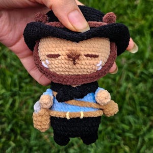 Puede incluir: Una pequeña figura de peluche tejida a ganchillo con cabeza color canela y ojos cerrados, que lleva un sombrero negro de ala ancha, una bufanda negra, una camisa azul claro y pantalones negros. Incluye un cinturón color canela con hebilla amarilla.