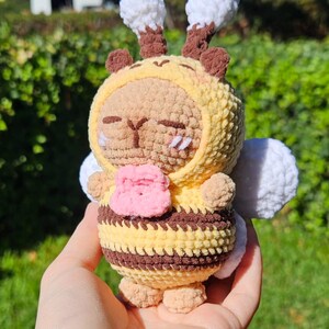 Puede incluir: Un pequeño peluche de ganchillo hecho a mano con forma de abeja. La figura presenta un cuerpo a rayas amarillas y marrones, una cara marrón claro con ojos bordados y una flor rosa en sus brazos. Tiene alas blancas y dos antenas con puntas blancas.
