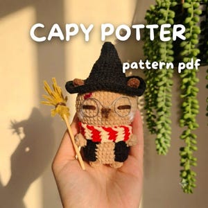 Puede incluir: Una figura amigurumi de ganchillo de un capibara con un sombrero negro puntiagudo, una bufanda de rayas rojas y blancas y gafas de alambre redondas, sosteniendo una pequeña escoba amarilla. El texto CAPY POTTER pattern pdf es visible.