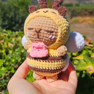 Puede incluir: Muñeca de abeja de ganchillo hecha a mano con cuerpo a rayas amarillas y marrones, chaqueta con capucha amarilla y alas blancas. La abeja tiene antenas marrones y boca rosa. La muñeca se sostiene en una mano sobre un fondo de follaje verde y cielo azul.