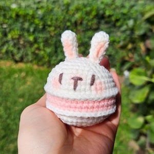 May include: A handmade crochet bunny toy in white and pink. The bunny has long ears with pink inner ears, a pink face, and a pink bottom. The bunny's face has a black 'T' shape. The toy is held in a hand.