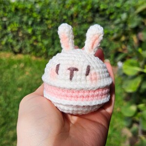 Puede incluir: Macaron de conejo de ganchillo hecho a mano en blanco y rosa. El conejito tiene orejas erguidas, orejas internas rosas y una cara cosida. El macaron tiene un relleno rosa.