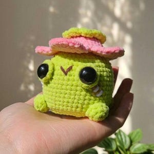 Puede incluir: Un pequeño juguete de peluche tejido a ganchillo de color verde lima, de forma redonda, con un sombrero rosa de ala ancha y borde amarillo. La figura tiene dos ojos circulares grandes de color negro brillante, una boca en V marrón y dos detalles blancos.