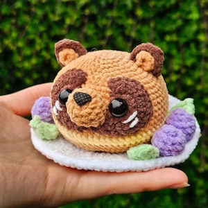 Puede incluir: Peluche de tanuki tejido a ganchillo con detalles marrones, ojos negros y un plato blanco. El tanuki está sentado en un plato blanco con detalles de ganchillo morados y verdes. El peluche está hecho a mano y tiene un diseño lindo y de dibujos animados.