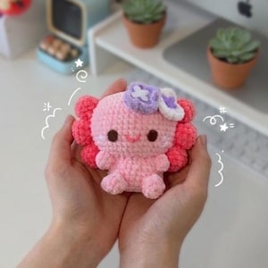 Puede incluir: Un pequeño peluche de axolote tejido a ganchillo a mano en tonos rosa claro y rosa oscuro. La criatura presenta ojos de color marrón oscuro, una sonrisa bordada y un accesorio de flor morada y blanca en la cabeza, con líneas de garabatos blancas.