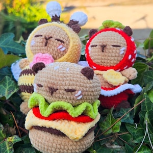 Puede incluir: Tres juguetes de peluche hechos a mano con ganchillo: una abeja, una fresa y una hamburguesa. La abeja es amarilla y marrón, la fresa es roja con puntos blancos y la hamburguesa tiene lechuga, tomate y queso. Los tres tienen los ojos cerrados.