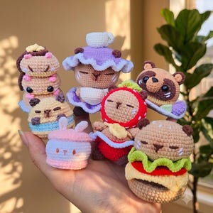 Puede incluir: Una colección de pequeñas figuras amigurumi de ganchillo con varios diseños, incluyendo una pila de criaturas color canela, una figura con sombrero morado, un personaje con temática de fresa, un mapache color canela, un objeto rosa con orejas de conejo y una hamburguesa.