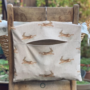 Könnte beinhalten: Eine beige Stoff-Aufbewahrungstasche mit einem sich wiederholenden Muster aus braunen Hasen in Laufposition. Die Tasche hat eine große Öffnung und hängt an einem Holzstuhl. Die Tasche ist für die Aufbewahrung bestimmt.