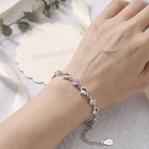 May include: A silver-toned chain bracelet featuring alternating open heart-shaped links and round, faceted pink stones. The bracelet includes a delicate extender chain with a small metal tag stamped with the text S925 at the end.