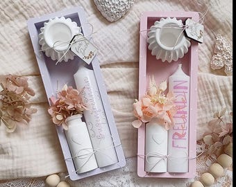 May include: Two decorative gift sets in rectangular boxes. One is lavender, the other pink. Each contains a white candle, a small vase, and a decorative flower arrangement. The candles have text on them. A white heart and wooden beads are also visible.