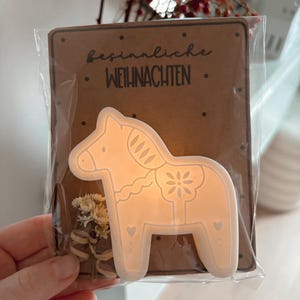 May include: A white Dala horse-shaped Christmas ornament with decorative cutouts, packaged with a brown card that reads "besinnliche Weihnachten". The ornament is in a clear plastic sleeve, with dried flowers.