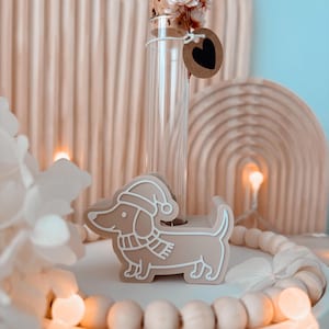 以下が含まれることがあります： サンタ帽とスカーフを身に着けたベージュ色のダックスフンドの置物が白いトレイの上にあります。置物の後ろには、コルク栓と小さなハート型のタグが付いた透明なガラス瓶があります。背景には、明るい色のアーチのデザインがあります。