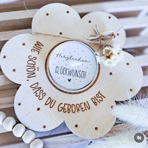 Puede incluir: Un portavelas de madera con forma de flor con el texto "Wie schön, dass du geboren bist" y "Herzlichen Glückwunsch" en él. El portavelas tiene una pequeña vela en el centro y un pequeño ramo de flores secas en la parte superior.